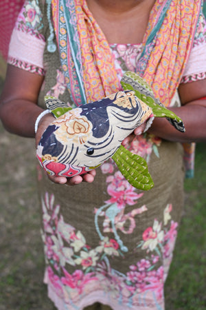 A soft, handmade stuffed whale toy made from colorful kantha fabric. The whale features colorful saree patterns in vibrant shades of red, blue, yellow, and green, with a rounded body, a curved tail, and a friendly face. The textured fabric adds a unique, artisanal touch to the toy, making it both a decorative and cuddly piece.